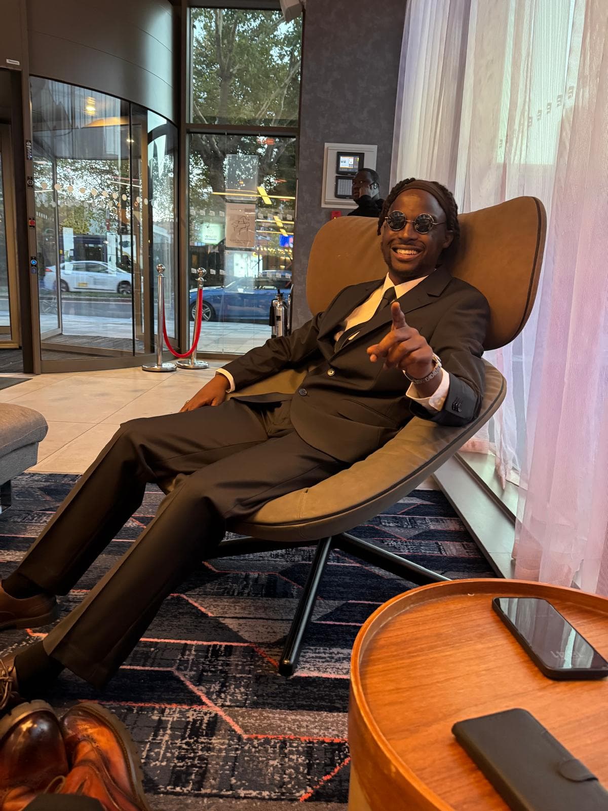 Emperor Roy seated in a hotel lobby, suited with sunglasses